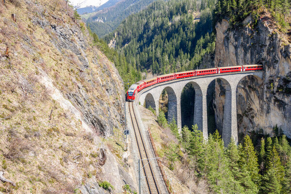 Красный электрический пассажирский поезд над изгибом каменного виадука в горах в солнечный весенний день. Landwasser Viaduct, Fillisur, Швейцария