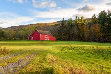 Sonbaharda günbatımında ormanlık dağ manzarasında çimenlik bir arazide geleneksel kırmızı Amerikan ahşap ahırı. Sonbahar renkleri. Catskill Dağları, New York, ABD.