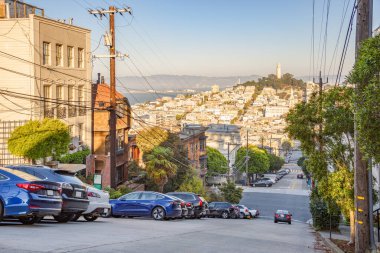 San Francisco 'da güneşli bir sonbahar gününde kaldırım kenarına park edilmiş binalarla dolu dik bir cadde. Coit kulesi uzaktan görülebilir. Kaliforniya, ABD.