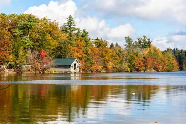 Sahildeki ahşap kayıkhane güneşli bir günde sonbahar renklerinin zirvesinde ormanla çevrili bir göl. Dublin Pond, Dublin, NH, ABD.