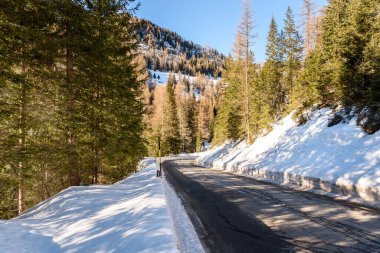 Açık bir kış gününde, karla kaplı ormanlık yamaçlar arasında uzanan düz bir dağ yolu.