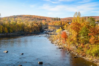 Kemerli yol köprüsü, güneşli bir günde sonbahar renklerinin zirvesindeki ormanlık bir arazide bir nehri geçiyor. Rumford, ME, ABD.