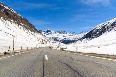 Empty mountain road overlooked by towering snowy peaks under blue sky in the Swiss Alps in early spring. Julier Pass, Switzerland. clipart