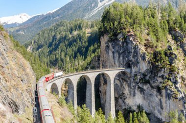 Yolcu treni, güneşli bir bahar gününde İsviçre Alpleri 'ndeki bir tünele giden taştan bir viyadük üzerinde beyaz bir elektrikli lokomotifle çekildi. Filisur, İsviçre.