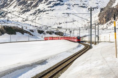Güneşli bir bahar günü İsviçre Alplerinde karlı yüksek bir dağ geçidinden geçen kırmızı yolcu treni. Bernina Geçidi, İsviçre.