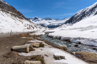 İsviçre Alpleri 'nin görkemli zirveleriyle çevrili karlı bir vadiden geçen bir yol boyunca akan akarsu açık bir bahar günü. Julier geçidi, İsviçre.