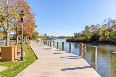 Açık bir sonbahar sabahında, bir nehir limanında ağaçlar, ahşap banklar ve lamba direkleriyle dolu boş bir tahta kaldırım. Demir atmış tekneler uzaktan görülebilir. Riverhead, Long Island, New York, ABD