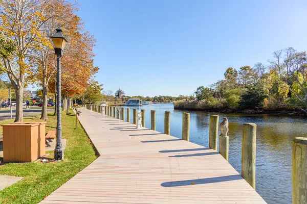 Açık bir sonbahar sabahında, bir nehir limanında ağaçlar, ahşap banklar ve lamba direkleriyle dolu boş bir tahta kaldırım. Demir atmış tekneler uzaktan görülebilir. Riverhead, Long Island, New York, ABD