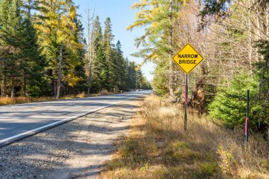 Açık bir sonbahar gününde dağlarda düz bir orman yolu boyunca dar bir köprüye karşı trafik işareti uyarısı. Grafton Notch, ME, ABD.