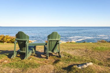 Boş tahta Adirondack sandalyeleri açık bir sonbahar gününde Maine kıyısındaki bir uçurumun tepesinde okyanusa bakıyor. Pemaquid Point, ME, ABD.