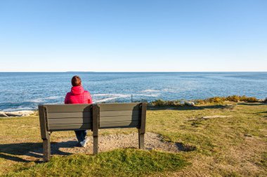 Açık bir sonbahar gününde uçurumun tepesindeki bankta tek başına oturmuş muhteşem deniz manzarasının tadını çıkaran bir kadın. Pemaquid Point, ME, ABD.