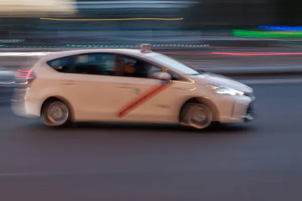 Madrid taksisinin hareketli görüntüsü alacakaranlıkta trafiğin yoğun olduğu bir caddede ilerliyor ve arka plan aydınlatma tarzıyla bulanıklaşıyor.