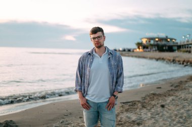 Deniz ya da okyanusun arka planında gözlüklü ve ekose gömlekli yakışıklı bir genç. Seyahat tatili. Denizde yürüyen bir adam tropikal mevsimin tadını çıkarır..