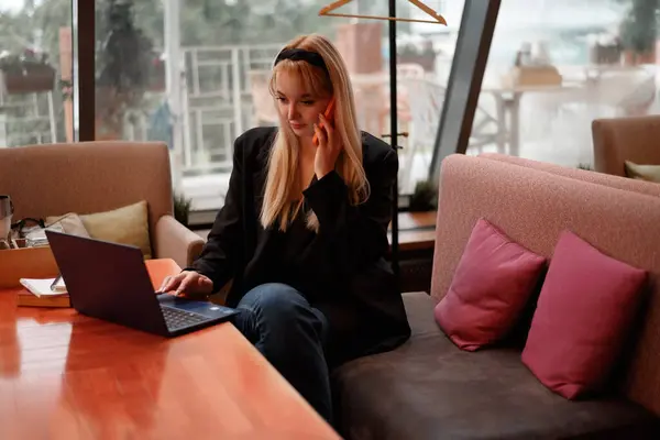 stock image Freelancer woman sitting in a coffee shop with a laptop on the table while making notes and talking on cellphone. Focused businesswoman in casual outfit drink coffee while working remotely in cafe