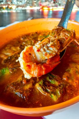 Bir kase baharatlı çorba ve kaşıkta karides, Tayland usulü yemek. Şehir manzarasını gösteren bir penceresi olan siyah bir tezgahın üzerinde, arka planda bir şehir silüeti olan