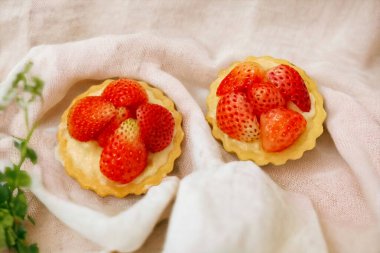 Açık kahverengi bir kumaşın üzerinde çilekli küçük pastalar ve arka planda yemyeşil bir çayır.