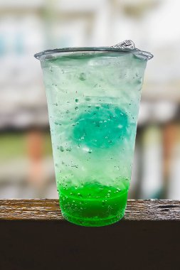 Plastik bardakta buzlu yeşil soda, ahşap masa ve bulanık arka plan.