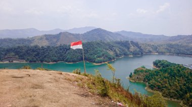 Dağda asılı duran kırmızı ve beyaz Endonezya bayrağı. Yeşil suyu olan diğer dağ sıralarını ve gölleri görebilirsiniz. Yeşil ve sarı ağaçlar, diğer bitkiler ve bulutlu mavi bir gökyüzü var..