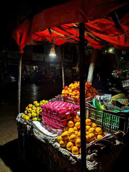 Hindistan 'da portakal, elma ve limon satan bir gece marketi. Meyve plastik kaplarda düzgün bir şekilde sergileniyor ve parlak bir ışık ekranı aydınlatıyor. Kabin kırmızı bir muşambayla kaplı.