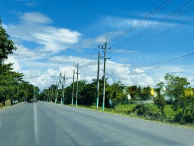 Geniş, asfaltlı bir yol, tüylü beyaz bulutlarla dolu açık mavi bir gökyüzünün altında dümdüz uzanır. Yol, kenarı boyunca uzanan elektrik hatlarıyla yeşil ağaçlar ve yemyeşil yapraklarla çevrilidir. Güneş ışıl ışıl parlıyor, sıcak bir parıltı yayıyor