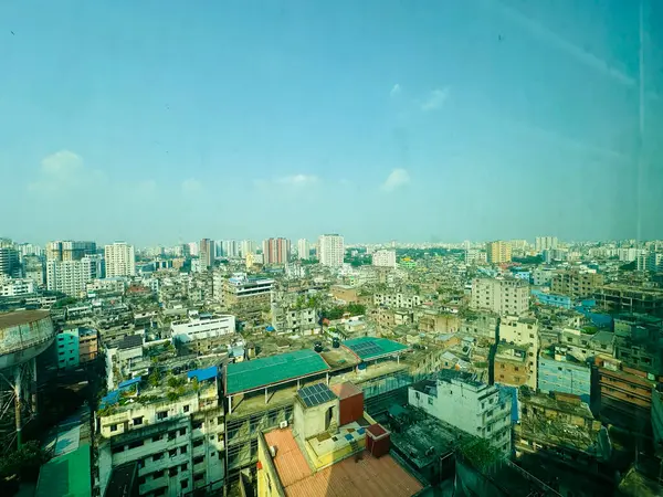 Güneşli bir günde Dhaka, Bangladeş 'in panoramik manzarası. Görüntü, farklı yüksekliklerde binaların bulunduğu yoğun bir kentsel ortamı gösteriyor. Gökyüzü açık ve mavi, birkaç beyaz bulutla. Binalar çoğunlukla kahverengi ve gri.