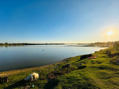 Güneşin batışının altın parıltısını yansıtan engin bir su kütlesinin yer aldığı sakin bir göl manzarası. Yemyeşil çimenler bankayı kaplıyor ve güzel bir zemin oluşturuyor. Açık mavi gökyüzü ufuk boyunca uzanır ve dinginliği arttırır.
