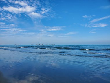 Sakin bir sahil sahnesi berrak gökyüzü ve yumuşak dalgalarla. Mavi bir okyanusta küçük balıkçı tekneleri yüzer..