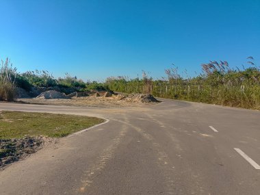 Uzak, güneşli bir yol kırsal bir alanda kıvrımlı, rüzgarda hışırdayan uzun otlarla çevrili. Kazılmış toprak yığınları ve son inşaat faaliyetlerine ait toprak parçaları. Temiz.