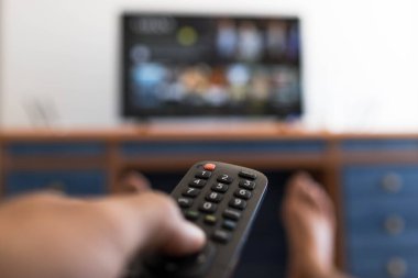 Uzaktan kumandayı elinde tutarken masada ayağı yere basan bir televizyon dizisi arıyor.