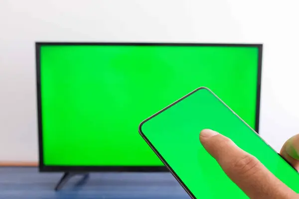 Elinde yeşil ekranlı, arka planda yeşil ekran televizyonu olan bir cep telefonu var.