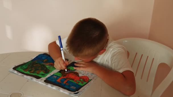8岁的男孩坐在桌旁 沉迷于着色书籍 每一页都讲述了他用生机勃勃的色调填充着色书籍的故事 录像陈列柜激发了孩子们对着色书籍的热情 — 图库视频影像