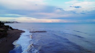 İnsansız hava aracı Akdeniz şehrinde süzülüyor, gün batımında sahil şeridi güzelliğini yakalıyor. Hava aracı görüntüleri: dalgalar kıyı şeridini okşadı, şehir zeminini. Günbatımı insansız hava aracı görüntüsü: sakin kıyı şeridi canlı şehir hayatıyla buluşuyor.
