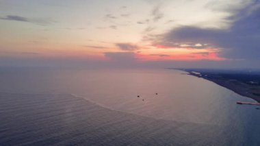 Bir İHA gün batımında muhteşem bir sahil şeridi görüntüsü yakalıyor. Gökyüzü parlak turuncu ve mor renklerle parlıyor, sakin okyanus suyuna yansıyor..