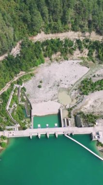 Altında turkuaz su bulunan yemyeşil bir ormandaki hidro barajın havadan görünüşü bu temiz enerji altyapısının huzur ve mühendislik harikasını gözler önüne seriyor.