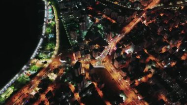Binalar ve sokak lambaları turuncu bir parıltı oluştururken, gece aydınlık şehir sokaklarından arabalar geçiyor. Canlı şehir manzarası, gece hayatının ve hareketli ulaşımın özünü yakalıyor.