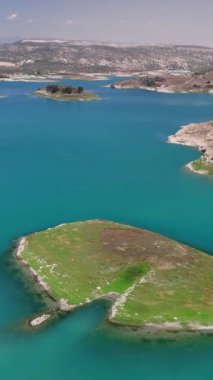 Turkuaz sular gün ışığı altında parıldarken, bir kamera adalarla süslenmiş ve yeşil tepelerle kucaklanmış huzurlu ve büyüleyici bir manzara yaratarak huzurlu bir gölün üzerinde süzülüyor.