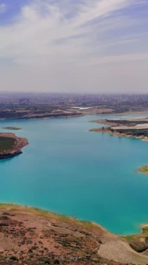 Bir gölün turkuaz suları bulutlarla benekli açık mavi bir gökyüzünün altında parıldıyor. Pürüzsüz bir kamera tepsisi, dingin manzarayı çevreleyen yemyeşil bitkileri ortaya çıkarır.