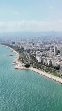 Sahil şeridinde berrak turkuaz sular boyunca uzanan mersin kenti bulunuyor. Binalar ve palmiye ağaçları kıyı boyunca uzanırken, uzak dağlar çarpıcı bir zemin oluşturuyor.
