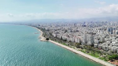 Sahil şeridinde berrak turkuaz sular boyunca uzanan mersin kenti bulunuyor. Binalar ve palmiye ağaçları kıyı boyunca uzanırken, uzak dağlar çarpıcı bir zemin oluşturuyor.