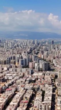 Şehir yayılımı parçalı bulutlu bir gökyüzünün altında uzanır ve uzak dağlar çarpıcı bir zemin oluşturur. Canlı bir şehir manzarası yerleşim bölgeleri ve altyapı karışımını sergiliyor