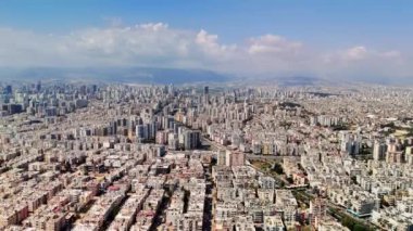 Şehir yayılımı parçalı bulutlu bir gökyüzünün altında uzanır ve uzak dağlar çarpıcı bir zemin oluşturur. Canlı bir şehir manzarası yerleşim bölgeleri ve altyapı karışımını sergiliyor