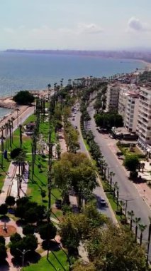 Türkçe kıyı şeridine çarpan turkuaz dalgalar, araçlar yolda ilerlerken canlı bir sahne yaratıyor. İnsanlar palmiye ağaçları ve çarpıcı mimariyle çevrili güzel bir günün tadını çıkarıyorlar.