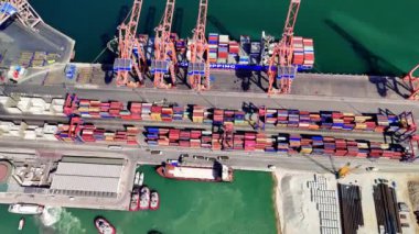 İşlek bir limanda yükleme ve boşaltma yapan vinçler, liman işçilerinin konteynırları taşıyan dinamik aktivitelerini sergiliyor. Lojistik ve deniz operasyonlarının canlı bir sahnesi güneşli gökyüzünün altında gözler önüne seriliyor.