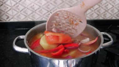 Şef, patatesleri, havuçları, soğanları ve kırmızı dolma biberleri karıştırarak lezzetli ve rahatlatıcı bir yemek hazırlıyor.