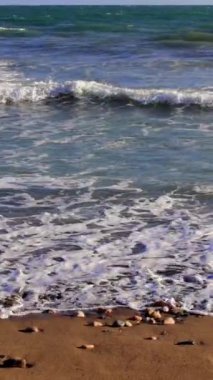 Turkuaz okyanus dalgaları yavaşça yuvarlanıyor ve kumlu bir çakıl taşı plajına çarpıyor. Huzurlu bir atmosfer yaratıyor. Sakin sular berrak mavi gökyüzü ile buluşur, sakin sahil manzarasını güçlendirir.