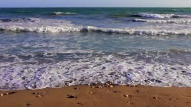 Turkuaz okyanus dalgaları yavaşça yuvarlanıyor ve kumlu bir çakıl taşı plajına çarpıyor. Huzurlu bir atmosfer yaratıyor. Sakin sular berrak mavi gökyüzü ile buluşur, sakin sahil manzarasını güçlendirir.