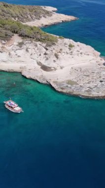 Berrak bir turkuaz koyda, yemyeşil bitki örtüsüyle süslenmiş kayalık bir kıyı şeridinin yanında küçük tekne çapaları. Turistler güneşli kayalıklarda dinlenir, dingin çevrenin tadını çıkarırlar.