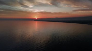 Canlı turuncu ve kırmızı renkler sakin bir denizin üzerinde gün batımında gökyüzünü doldurur. Güneş bir sahil şehrinin arkasına batar, suya yansıyarak huzurlu bir ortam yaratır.