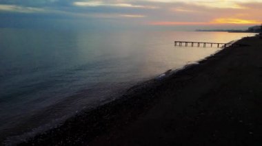 Sakin manzara, günbatımında kıyıya vuran deniz dalgalarını sergiliyor. Arka planda bir iskele ve güzel bulutlu bir gökyüzü var. Huzurlu bir kıyı manzarası açılır