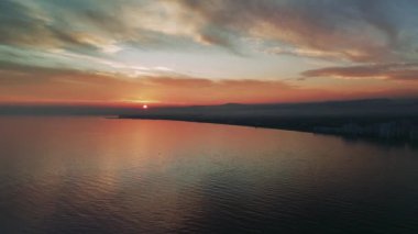 Akdeniz kıyı şehri üzerinde bulutlu bir akşam gökyüzünde canlı renkler bırakan gün batımı. Yansımalar suda dans eder, huzurlu ve resimsel bir sahne yaratır.
